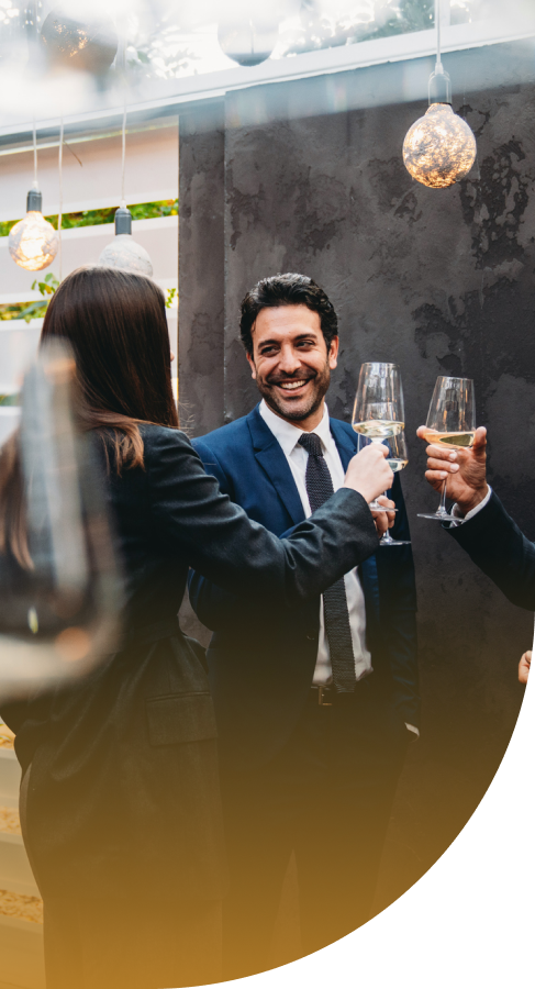 A man in a suit laughs while clinking champagne glasses with others at a festive gathering.